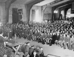 English: Rabbi Hershel Schaecter officiates at Shavuot services for Buchenwald survivors. Pictured in the audience are Robert Yehoshua Büchler (front row, shorts), Yisrael Meir Lau, and other notable individuals.